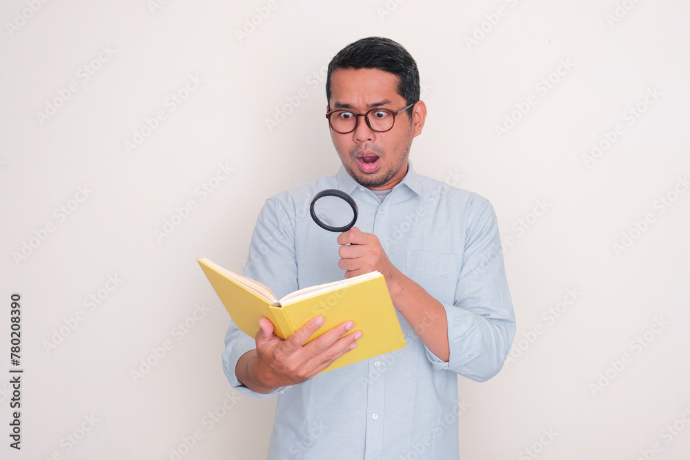 Adult Asian man reading a book through magnifying glass with shoccked face expression