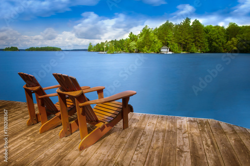 Two Adirondack chairs rest peacefully on a wooden dock, gazing out over the serene, blue lake. A single cottage, nestled among lush trees across the water, adds to the tranquil scenery.