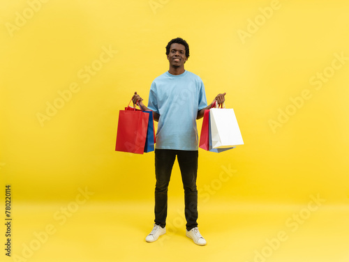 Young Man In Casual Wear Holding Shopping Bags Over Yellow Background. Concept Of Sales, Black Friday, Consumerism. Studio Shot With Copy Space. Trendy Colorful Shopper Lifestyle.