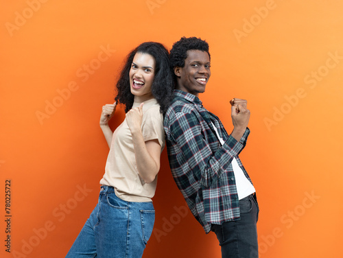 HAPPY MULTIETHNIC COUPLE CELEBRATING SUCCESS ON ORANGE Background. Diverse Joyful Partners, Energetic Poses, Modern Youth, Casual Style, Team Victory Concept. Stock Image For Commercial Use.
