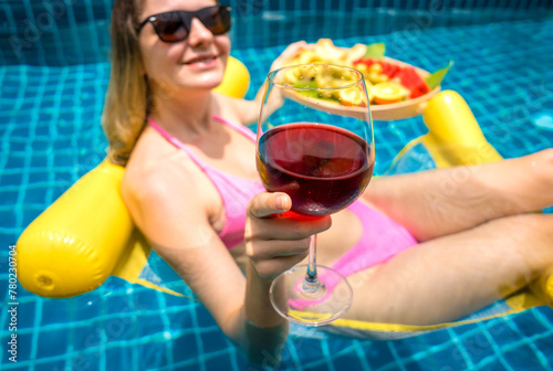 Beautiful woman lying on floating hammock in the swimming pool with wine and tray of fruits