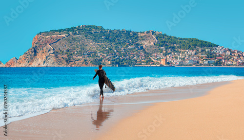 A male surfer walks on the beach with a surfboard in hand -  Landscape with marina and Red tower in Alanya peninsula, Antalya district, Turkey