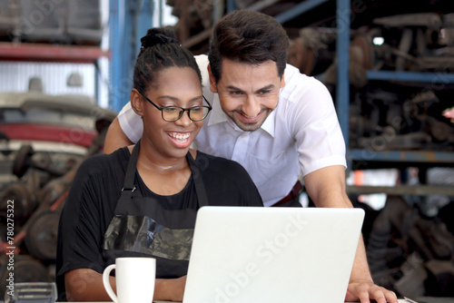 Happy harmony people work together at workplace, smiling African American woman working on laptop computer with colleague guy at auto spare parts store warehouse, surrounded by secondhand engine parts
