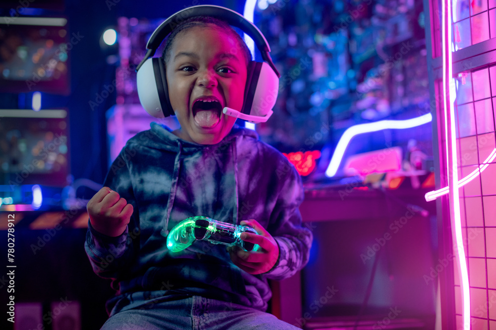 African child boy wear headphone excited playing video game, controlling characters with joystick at colorful neon light living room at night, E-sport streaming game online