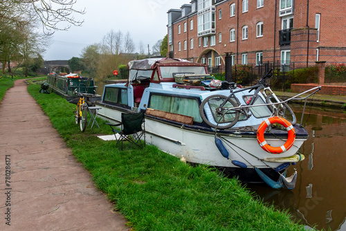 A canal house boat is moored by the towpath. There are bicycles parked outside. One is on the grass the other on the front of the boat.
