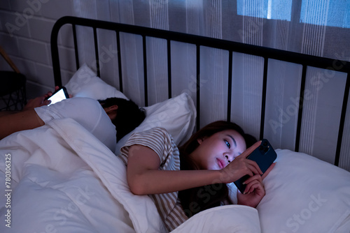 Asian couple lies back to back engrossed in their smartphones before sleep highlighting modern technology impact on intimacy and bedtime routine in a dark bedroom