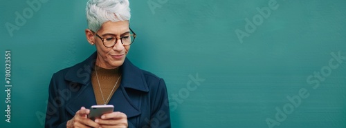 Photo of A mature businesswoman with short silver grey hair and glasses using her smartphone on green background, copy space for text. Web banner showing a woman smiling while looking at phone