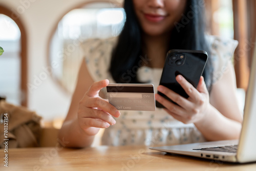 An Asian woman holding a credit card and a smartphone while sitting at a table in a cafe.