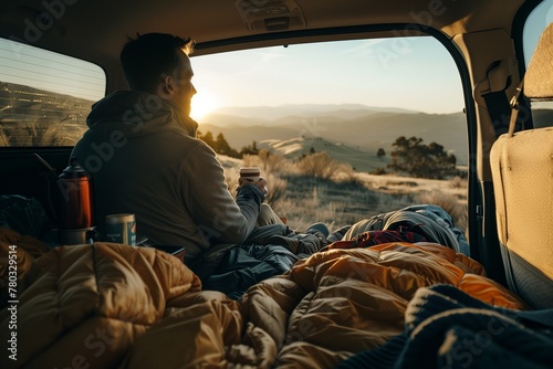 A man sitting in the trunk of his car, holding coffee and looking at mountains through an open window with blue blankets on him, camping gear inside, sunset light