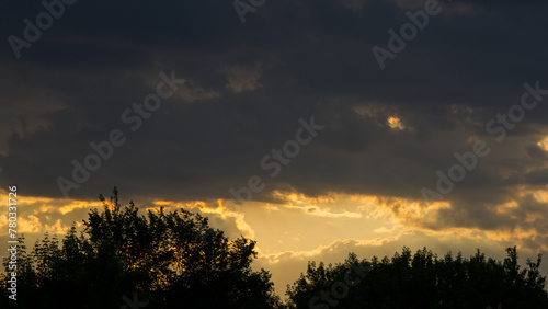 Dark clouds cover the horizon at sunset and dark silhouettes of trees.