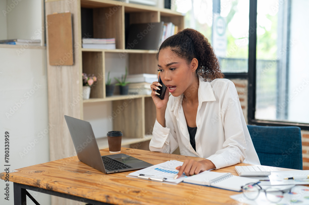 Young professional woman engaged in a phone conversation while working on her laptop at an organized office desk. Young professional woman engaged in a phone conversation while working on her laptop at an organized office desk.