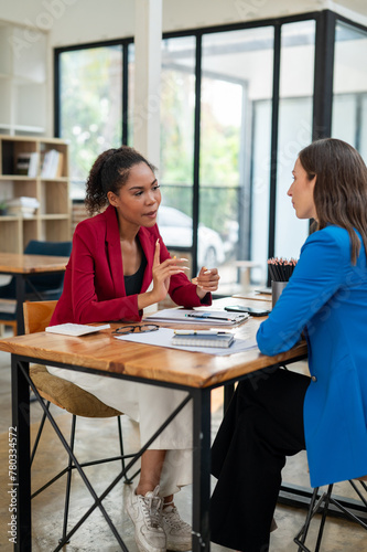 Two professional women in a lively discussion at an office desk, with one in a yellow blazer gesturing and the other showing surprise.