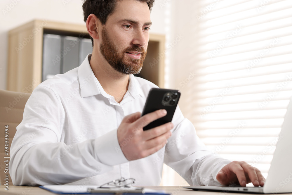 Man with smartphone using laptop at table in office