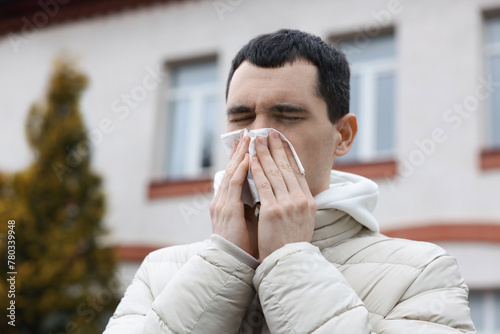 Sick young man with tissue blowing runny nose outdoors. Cold symptom