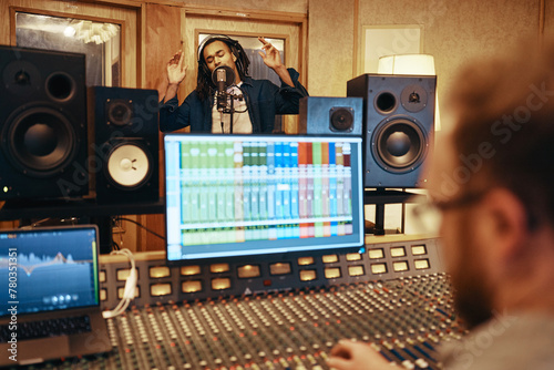 Producer adjusting sound levels during a studio recording session. A black charismatic singer stands and sings with arms raised and eyes closed, into a microphone photo
