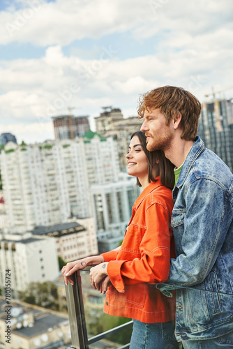 Фотография A man and a woman stand on a balcony, gazing at the city lights below, lost in t