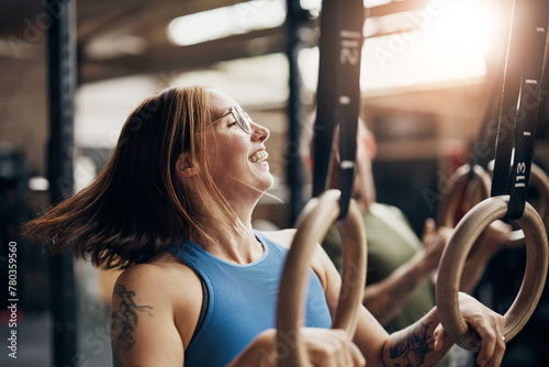 Smiling woman working out on rings during a gym exercise session. She has tattoos on her arms, and is wearing a blue tank top. Her friends are in the background. photo