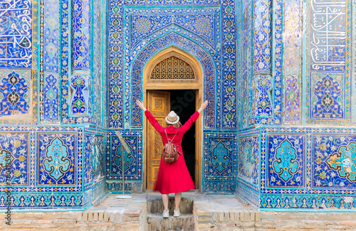 tourist woman in red dress clothes stands near an ancient architectural monument with wooden doors in oriental style with Islamic ornaments in Samarkand Uzbekistan Central Asia