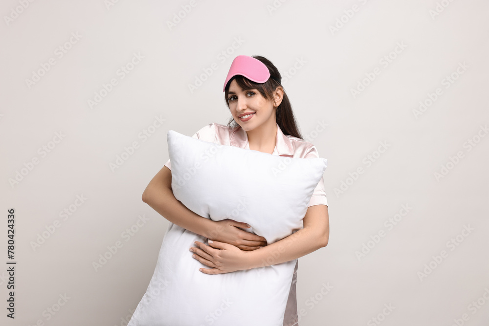 Happy woman in pyjama and sleep mask holding pillow on light grey background Happy woman in pyjama and sleep mask holding pillow on light grey background