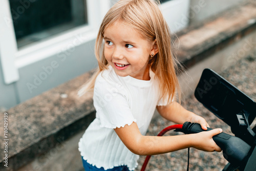 Smiling blond girl plugging charger in electric car at front yard photo
