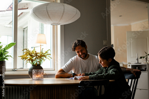 Smiling man sitting by son writing in book while doing homework at table photo