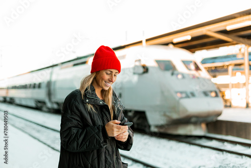 Smiling woman text messaging on mobile phone while standing at railroad station