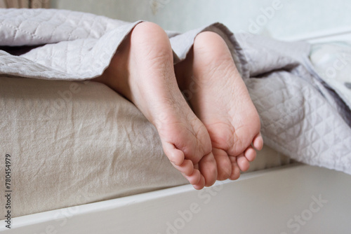 Feet under a light blanket on the bed, soft focus background © lisssbetha
