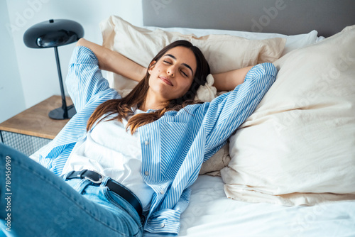 One serene young woman having relax laying on bed wearing casual clothes and smiling with closed eyes. People and travel enjoying hotel room. Morning indoor lazy leisure activity lady in relaxation photo
