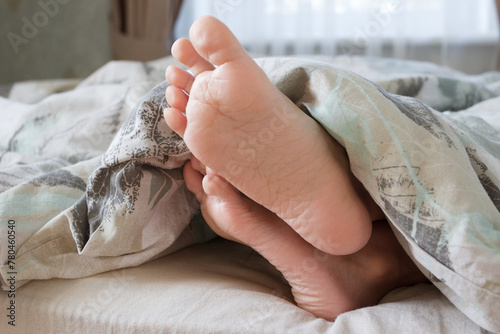 Feet under a light blanket on the bed, soft focus background © lisssbetha