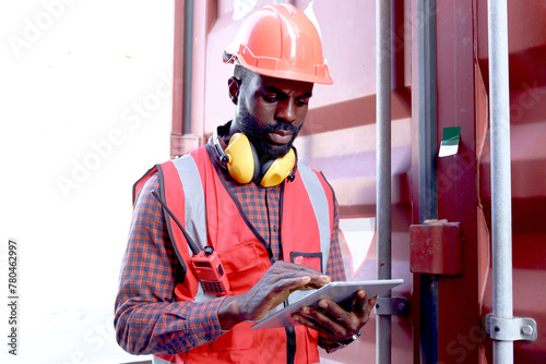 African American young engineer worker man wearing safety bright red vest and helmet, checking data from tablet at logistic cargo container shipping yard. A male worker working at workplace.