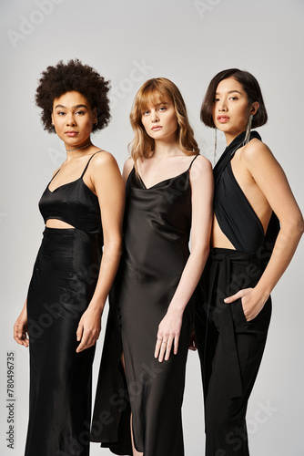 Three women of different ethnicities standing together in black dresses against a grey background, showcasing diversity and elegance.