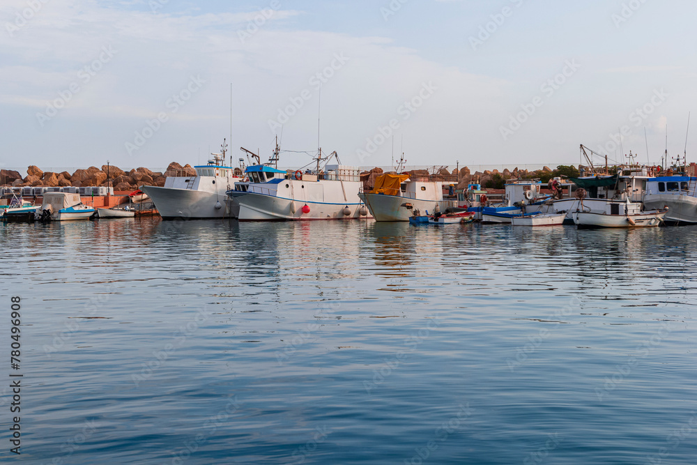 Fototapeta premium Stone pier in the marina of Tonnarella in Sicily. Fishing boats and dinghy boats on the water surface against the blue cloudy sky