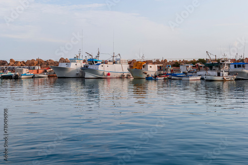 Stone pier in the marina of Tonnarella in Sicily. Fishing boats and dinghy boats on the water surface against the blue cloudy sky