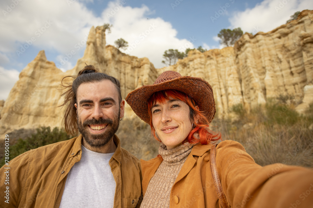 A man and a woman are posing for a picture in front of a mountain. The man is wearing a brown jacket and the woman is wearing a brown hat. Scene is cheerful and lighthearted, as the couple is smiling