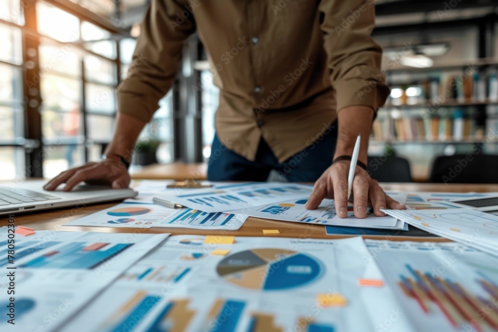 Businessman working on laptop while analyzing graphs and papers on table in office setting Businessman working on laptop while analyzing graphs and papers on table in office setting