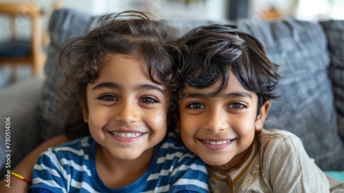 Two young children with dark hair smiling at the camera sitting close together on a gray couch with blurred background suggesting a cozy indoor setting.