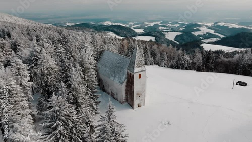 An aerial view of a quaint church nestled among a pristine snow-covered landscape, with dense coniferous forest in the foreground, Pohorje, Areh, Slovenija photo