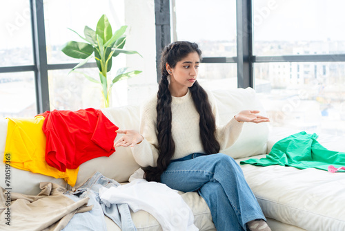Картината върху платно Distraught indian girl sits on a couch, looking indecisive as she struggles to choose what to wear