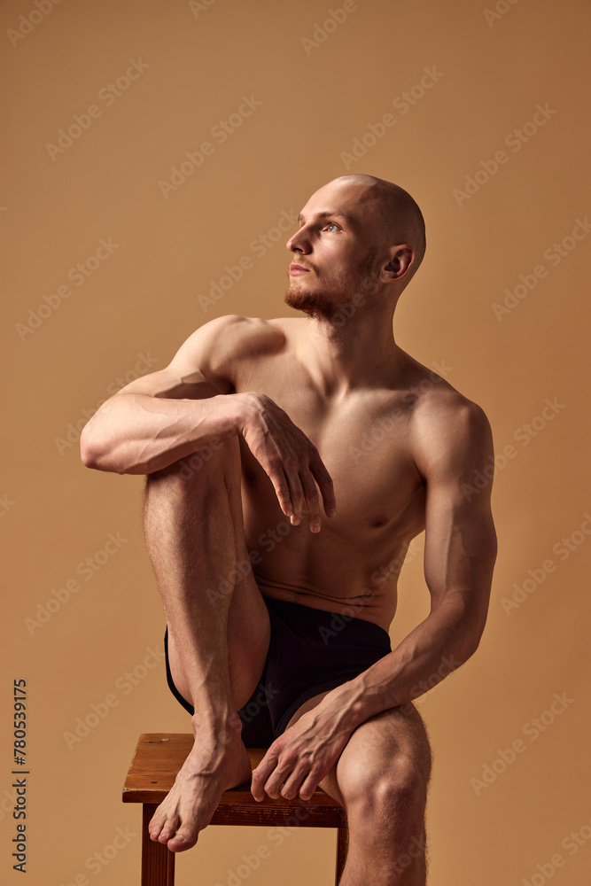Portrait of young man with athletic body posing in underwear on stool looking away against sandy studio background. Concept of men's health, self care, fashion and beauty, healthy lifestyle. Ad - obrazy, fototapety, plakaty 
