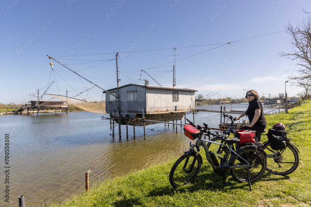 Woman cycles on an e-bike through the Po Delta in Northern Italy. In the background is a typical Italian pile dwelling, called Trabucco