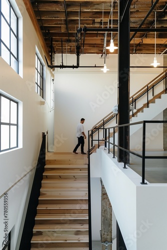 minimalist interior design  white walls with wooden beams on the ceiling  wood staircase  light brown stairs handrail  black metal balustrade  man walking up the stairwell  architectural photography