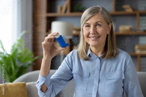 Smiling woman with blue inhaler in hand looking at camera while sitting in domestic atmosphere. Wrinkled female getting used to daily routine for health and wellness with chronically disease. photo
