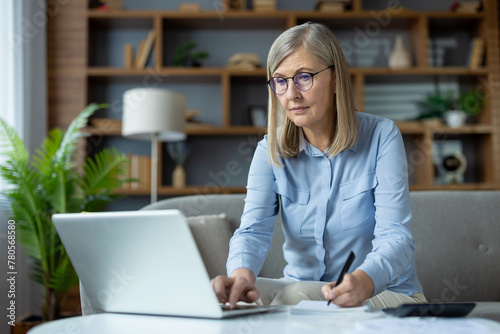 Canvas Print Stylish mature woman sitting on couch and using portable computer while writing in notebook at home