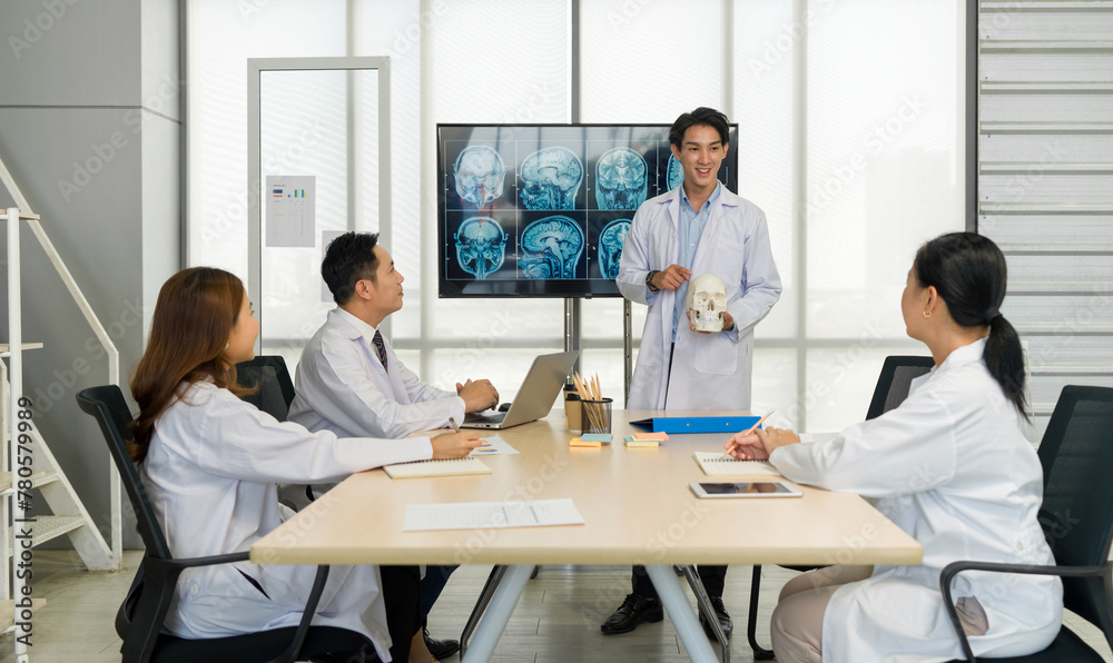 Young medical professional standing and presenting, holding a model of human skull and referencing image of brain scan displayed on a screen. The audience listening attentively with the presentation. Young medical professional standing and presenting, holding a model of human skull and referencing image of brain scan displayed on a screen. The audience listening attentively with the presentation.