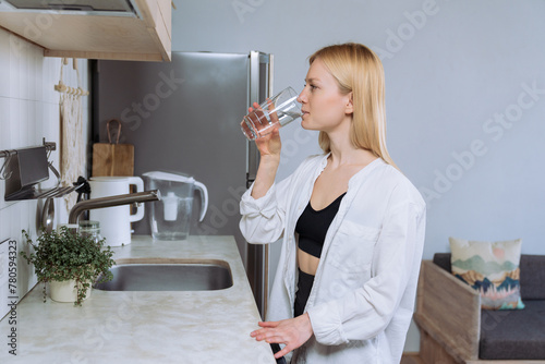A young woman drinks clean water from a glass in the kitchen.