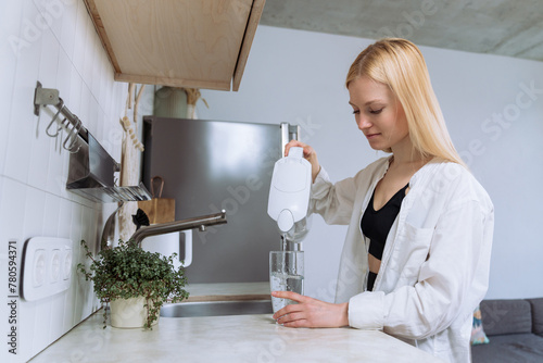 A young woman pours clean water into a glass in a bright and clean kitchen.