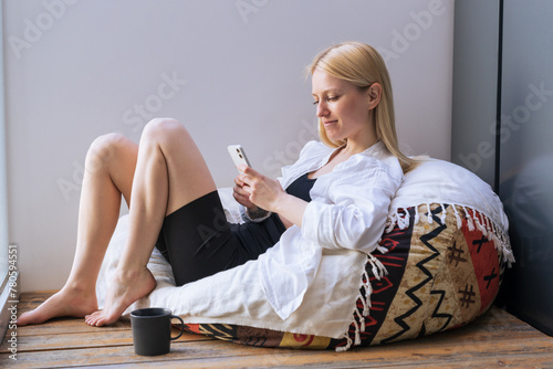 A young woman surfs the Internet from her phone and drinks coffee on the balcony.