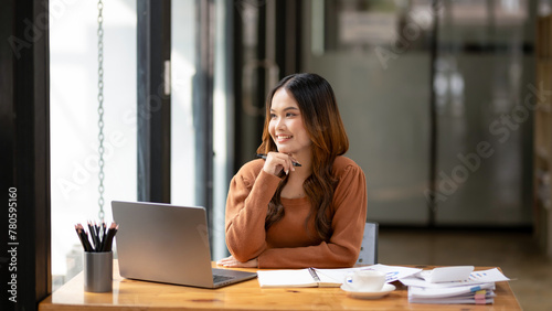 A focused young woman working intently at desk with a laptop and notebook in a bright, modern office setting.