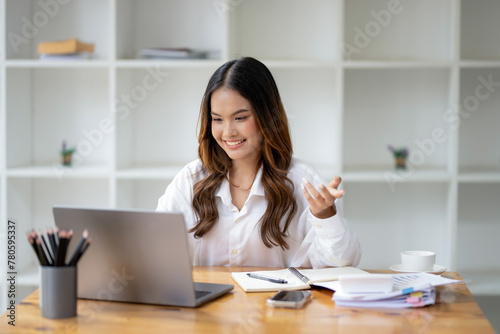 A young, smiling businesswoman confidently engages in a video conference call on her laptop in a modern office.
