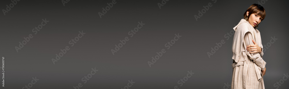 A young woman with short hair stands in trench coat against a serene gray background, exuding an aura of peace and purity. A young woman with short hair stands in trench coat against a serene gray background, exuding an aura of peace and purity.